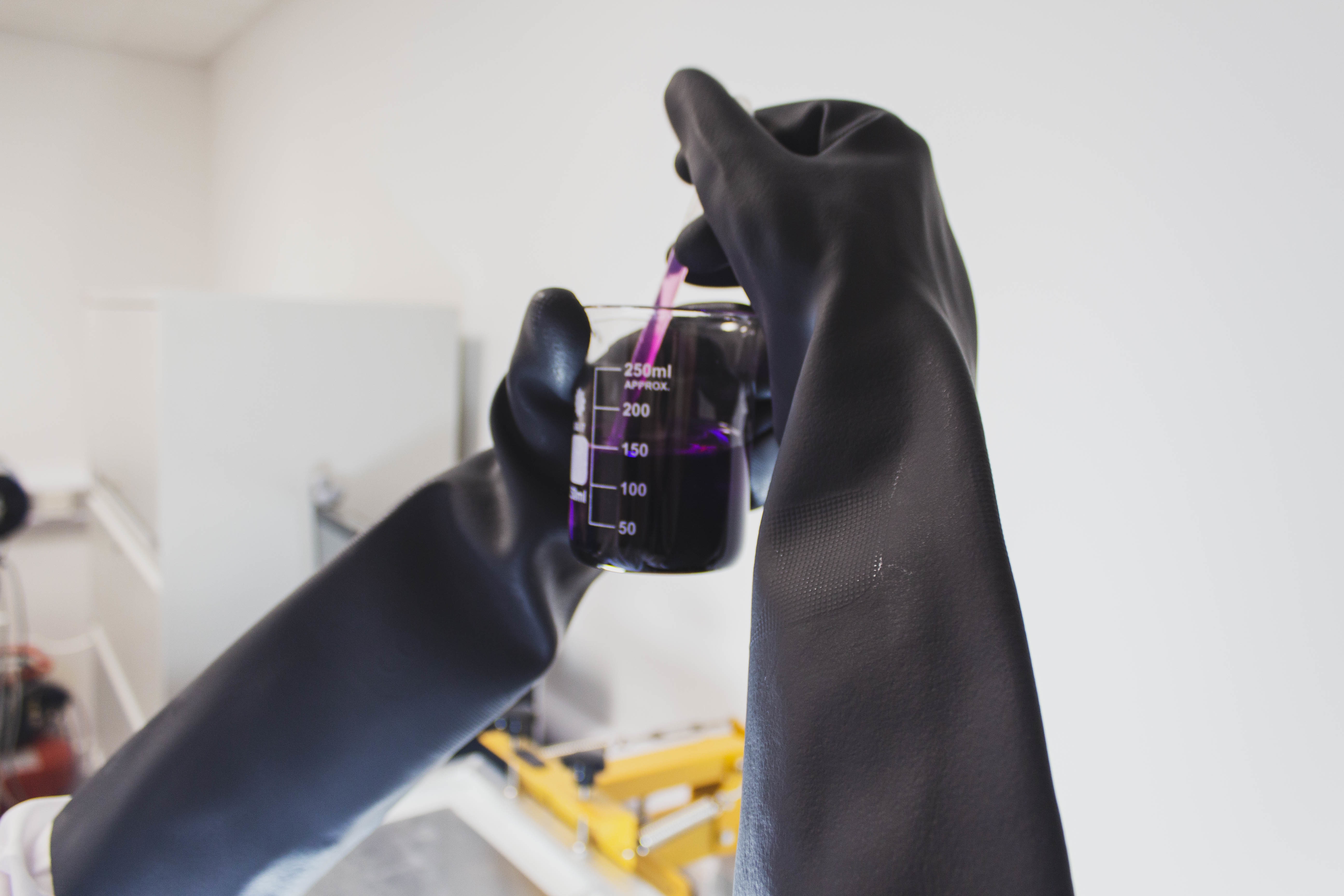 Black gloves handling a glass of chemicals.
